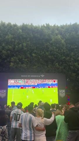 A wide shot captures a crowd of people gathered outdoors, facing a large screen. The screen displays a soccer match in progress, with players in green and white uniforms on a bright green field. The screen also features the logos of "BANK OF AMERICA" and "USA". The people in the foreground are partially obscured by the screen and are looking towards it. One person in the center is wearing a white top and has their arms around another person. To their left, someone wears a white and blue striped jersey with "MESSI 10" on the back. The background consists of lush green trees, creating a natural backdrop. The lighting suggests it is daytime or early evening. The overall scene depicts a public viewing of a sporting event.