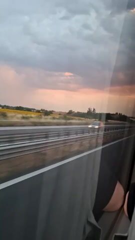 A view from inside a vehicle looking out a window at a highway and a cloudy sunset. The highway has multiple lanes and guardrails. The sky is filled with dark, heavy clouds, but there are patches of pink and orange light breaking through, indicating the sun is setting. The landscape beyond the highway appears dry and hilly, with some scattered trees and fields. The reflection of the inside of the vehicle is visible on the window, including part of a person's leg and black clothing. There are no distinct actions or interactions occurring in the video, it is a passive observation of the environment.