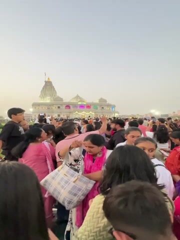 The video shows a large crowd of people gathered in front of a white, ornate building with multiple spires and domes. The building has the word "प्रेम मन्दिर" (Prem Mandir) displayed on the top in illuminated letters. Many people are taking pictures with their phones. The crowd is diverse, with people of different ages and genders. Some people are dressed in traditional Indian clothing. The sky is overcast.