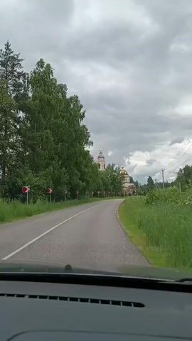 A dashcam view shows a car traveling on a road with a line of red and white directional signs indicating curves. Lush green trees line the left side of the road, and fields of grass are visible on the right. In the distance, two domed buildings, likely churches, with golden spires are visible. The sky is overcast with heavy clouds. The video shows a curved road, trees, grass, buildings, and a cloudy sky.