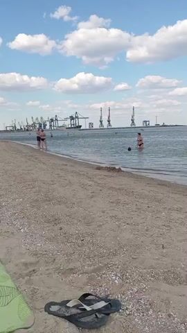 The video shows a beach scene with several people visible. In the foreground, on the sandy beach, there is a pair of flip-flops and a green object, possibly a towel or mat. The water is calm, and several people are in the water. To the left, two people are standing near the water's edge, facing away from the camera. In the middle distance, a person is standing in the water, and another person appears to be splashing or playing in the water. In the background, there is a body of water, and on the horizon, there is a port with several large cranes. The sky is blue with scattered white clouds.