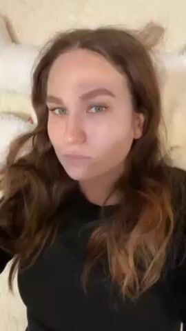 The video shows a woman with long, wavy, auburn hair wearing a black long-sleeved shirt. She is lying on a light-colored, possibly white, fluffy surface, presumably a bed or a blanket. She has fair skin, light blue or green eyes, and a nose piercing. Her gaze is directed towards the camera. She closes her eyes momentarily and then opens them again, maintaining a neutral expression. The camera remains still throughout the video.