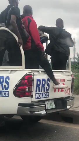 This video shows several police vehicles, including a pickup truck with "RRS POLICE" written on the side and a larger truck with red, white, and blue stripes and a flashing light on top. Some individuals are visible in the back of the pickup truck. In the background, there is a trash dump with some people present. The weather appears overcast.