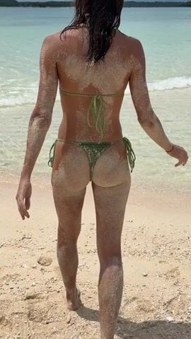 A woman in a green bikini walks on a sandy beach toward the ocean. She is covered in sand, especially on her back, arms, and legs. The water is clear and turquoise. She turns to face the camera, then bends down and puts her hands in the sand. She has dark hair and wears sunglasses.