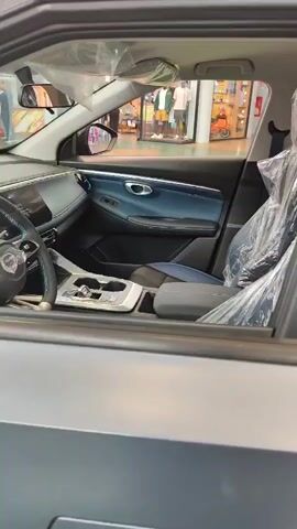 The video shows the interior of a car, viewed through the open driver's side window. The car appears to be new, with protective plastic coverings on the seats and steering wheel. The dashboard features a large screen and a modern steering wheel with controls. The center console has a gear shifter and cupholders. The interior is predominantly gray and blue. In the background, visible through the car's windows, is a brightly lit indoor environment that resembles a shopping mall or exhibition hall. Several people are casually walking around in the background.