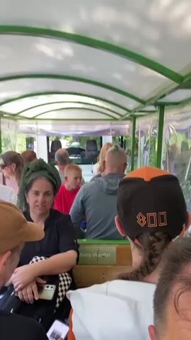 This media file captures a group of people riding on an open-air vehicle, likely a tour train or tram, through a lush, green environment. The vehicle has a green frame and a white roof, with clear plastic sheeting on the sides to offer some protection from the elements. Inside, several individuals are seated, facing forward.

In the foreground, a person wearing a brown baseball cap and a black and white checkered shirt is visible from the back. To their left, a woman with a green headscarf and a black top is seated, holding a smartphone. Beside her, a young boy in a bright red shirt is looking towards the front. Further back, other passengers are seated, some with their backs to the camera. The person in the immediate foreground has a braided ponytail and is wearing a dark baseball cap with orange trim and what appears to be the number "800" or similar graphic in orange on the back.

The scene appears to be outdoors, with greenery and trees visible through the plastic sides of the vehicle. The lighting suggests it is daytime, possibly late morning or afternoon, with bright, natural light filtering through the roof and the surrounding foliage. The overall mood seems relaxed and casual, consistent with a sightseeing or recreational activity. A small sign on the wooden interior of the vehicle reads "Many thanks!". The location context provided is Lipa Dobrska, Montenegro, which aligns with the natural, outdoor setting.