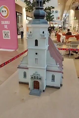 The video begins with a close-up of a miniature model of a white church with a dark steeple and a reddish-brown roof. The model is placed on a wooden surface within a shopping mall. In the background, several people are walking, and there are storefronts and a large pink display with the text "RIV CLUB" and a QR code.

The camera then pans to the right, showing the side of the church model and more of the mall environment. A sign with text and an image of a building is visible to the left. The scene shifts to show more of the shopping mall interior, including a coffee shop with tables and chairs, and other retail stores. Another miniature building model is visible in the foreground, displayed on a white platform with the word "MINIATURY" on it, and separated by stanchions. A man wearing a plaid shirt and shorts walks past the display. The video ends with a wider view of the shopping mall and the miniature displays.