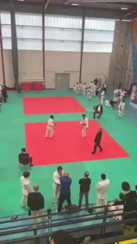 Two judo practitioners in white uniforms are engaged in a match on a red mat. One practitioner wears a green belt, the other a red belt. They circle each other, engaging in grips and attempting throws. The match takes place in a large indoor space with additional mats and spectators in the background. A man in a suit is seated on a chair near the mat, observing the match. Several people are standing near the edge of the mat, also observing. The environment is well-lit with natural light coming through large windows.