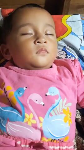 A baby is sleeping on a bed. The baby is wearing a pink t-shirt with flamingos on it and a pink and white striped skirt. The baby's eyes are closed and its arms are at its sides. The background is a bed with a wooden headboard and colorful sheets.