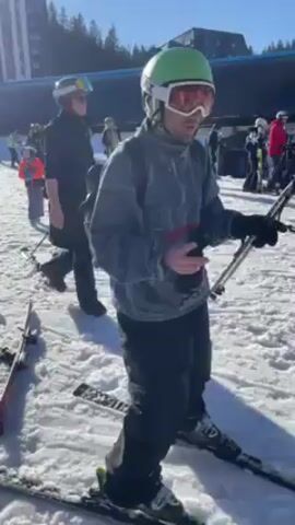 A man in ski gear is standing on a snowy surface and making hand gestures, smiling toward the camera. He is wearing a gray zip up, black ski pants, and a bright green helmet with ski goggles. He has ski poles in his hands and a black backpack on his back. Other people, also in ski gear, are standing nearby. Several pairs of skis are lying on the snow. In the background is a large building and a clear blue sky.