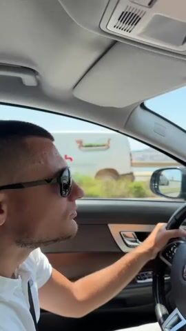 The video shows a man driving a Jaguar car. He is wearing sunglasses and a white polo shirt. The car is moving along a road next to a body of water with hills in the background. The weather appears to be sunny and clear. The man's hands are on the steering wheel and he is looking ahead. The interior of the car, including the dashboard and steering wheel, is visible.