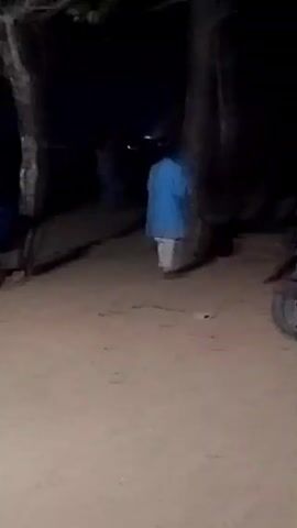 The video depicts a scene at night, where the ground appears to be sandy and there are several figures present. One individual is wearing a blue garment. There also appears to be a bicycle near the bottom-right corner of the frame. The area is sparsely illuminated, with visibility limited to the immediate surroundings. The environment includes trees that contribute to the dim lighting. The figures seem to be stationary, with no clear indication of activity beyond their presence in the area. The overall ambiance suggests a rural or undeveloped location at night.
