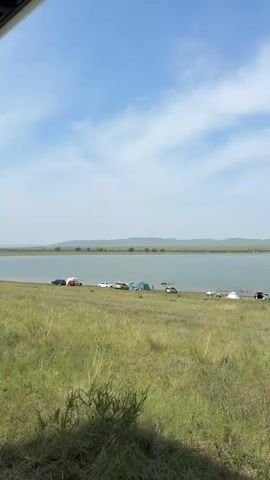 The video shows a daytime scene at a lake or reservoir. In the foreground, there is a grassy bank sloping down to the water. Several cars are parked on the grass near the water's edge, some with their trunks open. There are also a few tents set up. In the water, several people are swimming or wading. In the background, there is a wide expanse of water with a distant shoreline marked by a line of trees and low, rolling hills under a partly cloudy sky. The overall impression is of a relaxed outdoor recreation area.