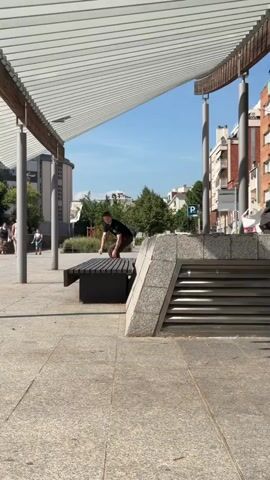 A young person on a skateboard jumps onto a park bench, then off the bench and out of frame. The skateboarder is wearing a black t-shirt, brown pants, and white shoes. They are in a park-like setting with a paved walkway, a bench, trees, and buildings in the background. The sky is blue and the sun is shining.