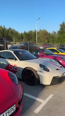 The video displays a line of vibrant Porsches parked in a lot. The cars are all different colors, including white with pink racing stripes, bright pink, yellow, dark grey, and red. The scene is outdoors, with a clear blue sky and lush green trees in the background. A tall light post is also visible. The cars appear to be in a parking lot, possibly at a car meet or gathering. The lighting suggests it is daytime, likely late afternoon or early evening, given the warm glow on the cars. There are no people visible in the frame, and the focus is entirely on the collection of sports cars. The overall impression is one of luxury and automotive passion. One car has "PORSCHE" written in pink on its hood, and another has "96" in a red circle on its side. License plates are partially visible on some of the cars, with text such as "CB 1800 XT" and "CB 1110 XP" discernible. The setting appears to be Kerenite, Bulgaria, as indicated by the license plate format.