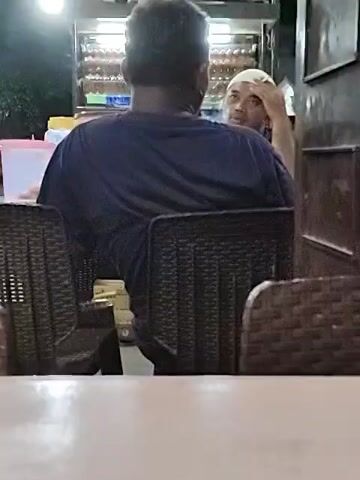 Two men are sitting at a table outside a food stall at night. The man facing the camera is wearing a white skullcap and resting his hand on his head. The other man is facing away from the camera. The food stall behind them has rows of food items on display. There are black wicker chairs around the table.