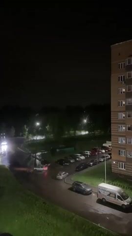 The video shows an urban night scene during a storm. A street runs through a residential area, lined with parked cars. Streetlights illuminate the wet pavement, reflecting headlights of moving vehicles. A tall apartment building stands on the right. In the background, there is a line of trees against the night sky, which is punctuated by flashes of lightning.