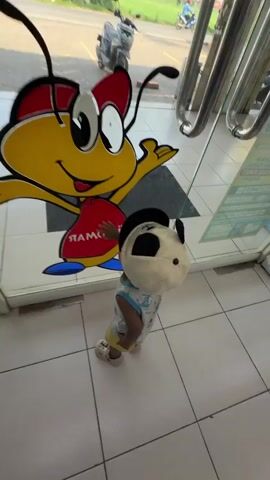 A young child wearing a panda mask and a light-colored tank top with a blue and white pattern, yellow shorts, and white sneakers stands in front of a glass door. The child's left hand is touching a cartoon bee sticker on the door. The cartoon bee is yellow with red wings and antennae, and it has a big smile. It is wearing a red bow and blue shoes, and its body is red with the word "RAMOS" written on it in white. The floor inside the building is tiled in white and gray squares. Outside, through the glass, a motorcycle is visible, as are other signs and buildings. The lighting suggests it is daytime.
