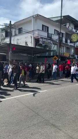 The video shows a large group of people walking across a street in a city. Many of them are wearing red shirts and hats, and a few are carrying red flags. The buildings on either side of the street are mostly two- and three-story structures with shops and businesses on the ground floor. The sky is partly cloudy. The people are walking in the same direction, some together, some separated. The overall impression is that of a march or demonstration.