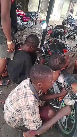 The video shows four young men working on a motorcycle. One is wearing a black shirt, another is wearing a plaid shirt, and a third is wearing a brown tank top. The fourth is standing up in a basketball jersey. They are squatting around the front of the motorcycle, appearing to be installing or adjusting something near the headlight. The ground is dirt and there are other motorcycles parked in the background.
