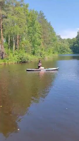 A person sits cross-legged on a stand-up paddleboard in the middle of a body of water. The person is wearing a dark t-shirt, a dark hat, and sunglasses. The paddleboard is white with colorful designs on it. The water is dark and calm with ripples reflecting the trees and sky. In the background, there is a shoreline covered in lush green trees and foliage. The sky is clear and blue. The person appears to be holding something small in their hands.