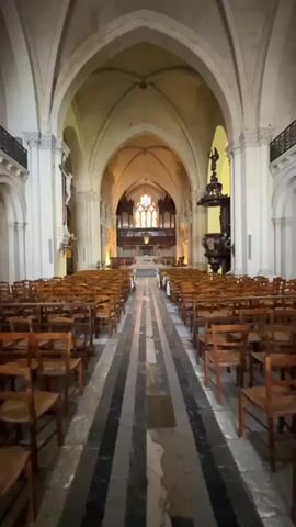 The video shows the interior of a large, empty church. The perspective is from the back of the nave, looking towards the altar. Rows of wooden chairs are neatly arranged on either side of a central aisle, which is paved with dark and light striped stone. The church has high, vaulted ceilings with arched supports. A pipe organ is visible at the far end, above the altar area, which is decorated with stained glass windows. To the right of the aisle, a dark wooden pulpit stands prominently. The overall lighting is dim, with some light filtering in from the windows and interior fixtures. There are no people visible in the video. The scene is still and quiet, suggesting an unoccupied space.