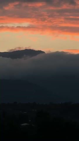 The video shows a view of a mountain range at sunset. The sky is filled with orange and pink clouds. The mountains are silhouetted against the sky, and there are some trees in the foreground. The overall mood of the video is peaceful and serene.