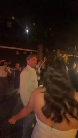 The video captures a crowded dance floor in a dimly lit nightclub. In the foreground, a man in a white collared shirt and light blue pants is dancing, facing away from the camera and towards another person whose back is to the viewer. Several other people are dancing around them, with their arms raised and bodies in motion. The environment is characterized by flashing lights, including vibrant red and blue hues, which illuminate the dancers and the space. The background shows a ceiling with lighting fixtures and some indistinct structural elements of the club. The overall atmosphere appears energetic and celebratory.