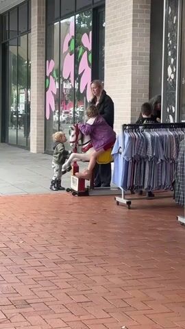 A young girl, wearing a purple jacket and red shorts, is sitting on a stationary rocking horse toy in front of a store. Her feet are bare. She is being assisted by a woman, likely her mother, who is standing behind her, holding her by the shoulders. Another young boy, wearing a grey jacket and patterned pants, stands to the left of the rocking horse, looking at the girl. A third child, a boy, stands to the right of the woman, looking forward. In the background, racks of clothing are displayed outside the store, and the store's facade features large windows with decorative pink floral designs. The ground is paved with reddish-brown bricks. The scene appears to be a public walkway in front of a retail establishment.