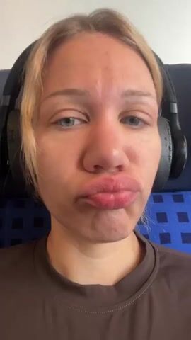 A person is sitting in a seat, possibly on a train or plane, as indicated by the blue and black patterned upholstery visible behind them and the presence of headphones. The person is looking directly at the camera and makes a kissing gesture before opening their mouth and pointing to a tooth with a small metallic object on it, possibly a dental ornament or piercing. The person is wearing a brown shirt. The lighting appears to be from an overhead source.