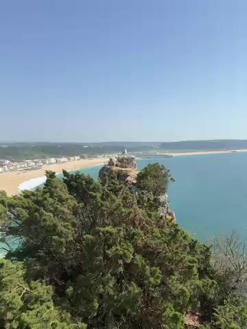 This video captures a panoramic view of a coastal landscape on a clear, sunny day. The scene is dominated by a long, golden sandy beach curving along the coastline, bordered by the turquoise waters of the ocean where white waves gently break.

In the foreground, lush green vegetation covers steep cliffs, including dense bushes, trees, and a distinctive agave plant growing on the rocky terrain. A prominent rock formation stands out, topped with a small white statue or structure, acting as a focal point.

As the camera slowly pans from right to left, it reveals a bustling town with numerous white and light-colored buildings nestled against the beach, stretching far into the distance. The bright sunlight illuminates the entire scene, creating clear visibility under a vast blue sky with only a few wispy clouds. The overall impression is one of a picturesque and serene coastal vista, likely overlooking Nazaré, Portugal, given the distinctive features. No people are explicitly visible in the immediate foreground, but the town and beach suggest human activity.