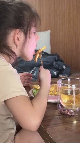 A young girl with brown hair and bangs, wearing a light-colored t-shirt, is sitting at a wooden table and eating. She is holding a fork with a piece of cooked food on it, and is taking a bite. On the table is a pink plate with more of the food and some sliced cucumbers, and a double-walled glass filled with a light-colored liquid. A grey blanket with star patterns is draped over the back of the booth seating behind her. Loud, upbeat music plays in the background.
