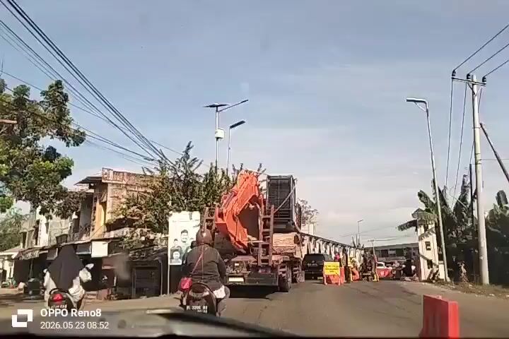 The video shows a street scene in what appears to be a small town or suburban area. A large orange excavator is being transported on a flatbed truck. There are workers in orange vests standing near orange traffic barriers, suggesting road work or a temporary obstruction. A motorcyclist is in front of the truck, moving in the same direction. There are buildings, trees, and utility poles lining the street. The weather is clear and sunny. The video is shot from the perspective of a vehicle following behind. The camera details include a date and timestamp from an OPPO Reno8 phone.