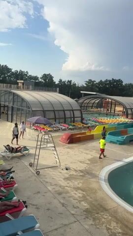 The video shows an outdoor water park on a cloudy day. In the foreground, there is a swimming pool with several people in it. A few people are lying on lounge chairs around the pool. There are some large animal sculptures in the shallow end of the pool. Behind the pool are several colorful water slides, some of which are covered by a transparent roof. There are people on the slides and in the water. In the background, there are trees and buildings. The overall impression is one of a busy and fun water park.