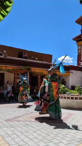 The video captures an outdoor cultural performance under a bright blue sky with scattered clouds. Two individuals, adorned in elaborate, colorful costumes and masks, are engaged in a dance. The costumes appear to be traditional, featuring vibrant green robes with intricate patterns and embellishments in red, yellow, and black. Both dancers wear masks that resemble animal heads, possibly deer, with prominent antlers. One dancer's mask also features blue accents around the head. The dancers are performing on a paved courtyard, with a building visible in the background. The building has multiple levels with balconies and windows, and people are observing the performance from the balconies and seated on the ground. The overall atmosphere suggests a festive or ceremonial event.