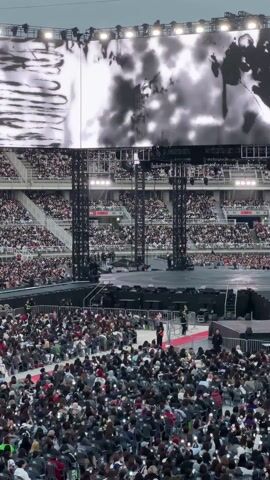 A large stadium is packed with people, with the camera angled from a high vantage point within the crowd. The audience is seated in folding chairs, holding up phones and recording the stage. A large screen displays visual imagery above the stage. Metal rigging supports the screen and stage lighting. The stadium seating rises in tiers, filled with more spectators. The overall atmosphere suggests a concert or performance event.