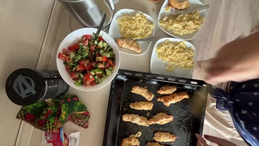 The video shows a top-down view of a meal laid out on a wooden table. The meal consists of a salad in a white bowl, pasta on three white plates, and chicken tenders on a black baking sheet. A person's hands are visible, using a fork to pick up a chicken tender from the baking sheet and place it on a plate. Another person's hands are visible in the background, also interacting with the food. A black supplement container is on the left side of the table. A patterned napkin is also visible. The lighting suggests an indoor environment.