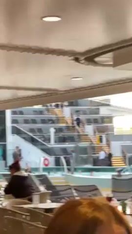 The video shows the outside of a cruise ship from an area under a canopy. There is a swimming pool with people in it and deck chairs nearby. A fair-skinned person wearing a light-colored button-down shirt suddenly comes into frame and excitedly walks forward. The camera follows the person as they continue walking through an indoor area on the ship.
