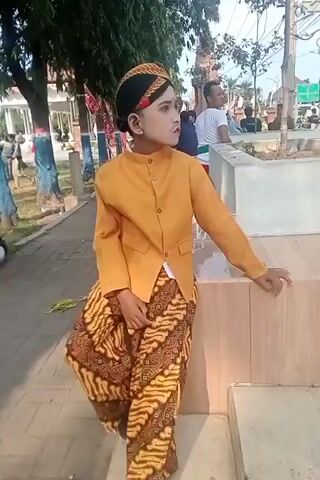 A young person in traditional Indonesian attire poses outdoors. They are wearing a mustard-colored jacket over a white undershirt and a patterned batik sarong. A traditional Javanese headdress, known as a blangkon, is on their head. The person is leaning against a low wall, with trees and other people in the background. The ground is paved with bricks. The lighting suggests it is daytime.