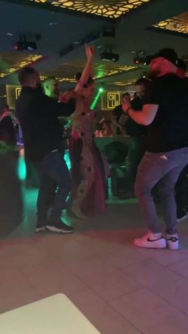 The video shows a belly dancer performing in a dimly lit club setting, surrounded by onlookers. The dancer is wearing a sparkly, elaborate costume with floral embellishments and a long, flowing skirt. She moves her arms and body rhythmically to the music. Several people are standing around her, some recording with their phones or clapping, while others interact directly with the dancer, including one man who briefly embraces her. The club has a modern decor with a lit-up sign that reads "CHI LAI" visible in the background. The dance floor is lit with green and purple lights.
