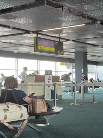 The video shows an airport gate area bustling with passengers and airline staff. In the foreground, a person is seated, resting with their feet up, holding a brown paper bag and a light-colored handbag. Ahead of them, a boarding counter is visible, where several people are standing, including two airline staff members and travelers with luggage.

A prominent digital display board overhead shows flight information: flight number JU 4684, destination Тиват (Tivat), scheduled departure at 17:00, and status "Идите на излаз" (Go to the gate) for gate C.13. A small timestamp in the corner of the display reads 16:45.

Next to the boarding counter stands an AirSERBIA sign detailing baggage allowances for "Ekonomska klasa" (Economy Class) and "Biznis klasa" (Business Class), illustrating permitted carry-on bags and personal items. The personal item has a maximum weight limit of 4kg.

The scene is brightly lit by natural light streaming through large windows, revealing an overcast sky outside. The overall atmosphere is that of a typical airport gate as passengers await their flight.