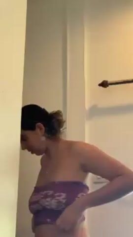 A woman in a purple strapless top opens curtains in a bright room with white walls. The curtains are two-toned, brown and tan.  A white fan is also visible.