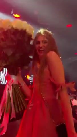 A woman with long, light brown hair is holding a large bouquet of white roses. She is wearing a pink strapless dress, and she is on a stage with red and blue lights. She is smiling and appears to be happy. The background is blurry, but it looks like there are other people on the stage and in the audience. The video seems to be taken from a slightly low angle, making the woman and bouquet appear larger.