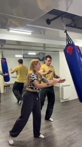 A group of people are in a room with a mirrored wall and a punching bag. A woman with long blonde hair is holding a blue punching bag by the top strap, while a man in a yellow t-shirt stands next to her. A boy in a patterned t-shirt is facing them. The woman lets go of the punching bag, and it swings. The man in the yellow shirt laughs. In the reflection, two other people can be seen.