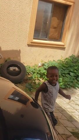 The video shows a young boy looking out from behind a black car parked in a residential area. The boy is smiling. The scene appears to be outdoors, possibly a courtyard or driveway, with a building made of pale yellow stucco. There is greenery, including a small bush and what looks like a palm tree trunk, visible in the background. Laundry is hanging on a line against the wall of the building. A black water barrel and a white bathtub are also visible. The ground is paved with hexagonal tiles. The lighting suggests it is daytime, with shadows cast on the ground. The overall atmosphere seems casual and domestic. There is no clear indication of the specific time of day or weather conditions beyond it being daylight.