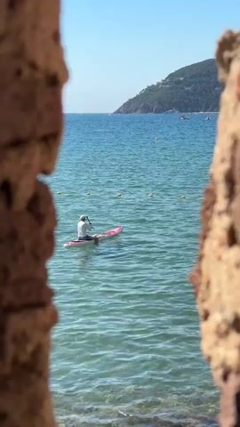 A person is paddleboarding in the ocean. The person is wearing a white shirt and a hat. They are holding a paddle and are in the middle of the ocean. The water is blue and clear, and there are some yellow buoys in the water. In the background, there is a hill covered in trees and a blue sky. The foreground of the video is blurred, showing parts of a brick wall.