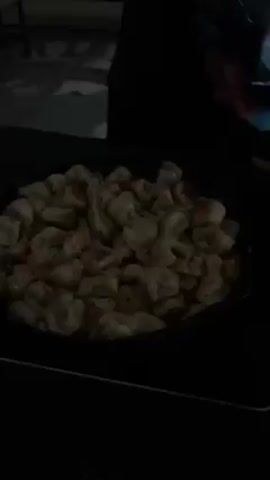 A person adds shredded cheese to a pan full of gnocchi. The pan is on a black stove. The background is dark.