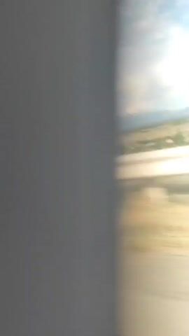 The video shows a view from a moving vehicle, looking out a window. A blue and white guardrail is visible on the right side of the frame, followed by a dirt road, grassy areas, and distant mountains under a partly cloudy sky. Utility poles and power lines also appear. The camera pans to the right, revealing more of the landscape.