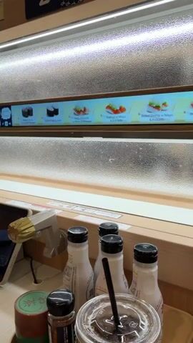 The video captures an interior view of a modern kaiten-zushi (conveyor belt sushi) restaurant, likely in Osaka, Japan. In the foreground, a wooden tabletop holds several items: four white bottles with black caps, a clear plastic cup with a straw, a small green container, and a small black shaker bottle, presumably for condiments or spices. A tablet device is partially visible on the left.

Above the table, two conveyor belt tracks are visible, each protected by a textured, translucent barrier. Above the lower belt, a digital screen displays various sushi menu items, including "Boiled Crab & Crab Butter Tokan," "Fatty Abalone w/Mayo & Avocado," "Salmon w/Mayo & Avocado," "Boiled Shrimp w/Mayo & Avocado," and "Prosciutto w/Mayo & Avocado."

An automated delivery is in progress on the upper conveyor belt. The screen associated with this belt shows a visual of sushi and the text "Large Silver Salmon 2 pcs," indicating that this specific order is being delivered to the customer. The restaurant appears well-lit and clean.