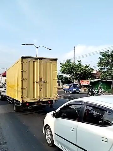 The video shows a street scene with several trucks and cars. A large yellow truck with a container is in the foreground. A red and white tanker truck with the logo "PERTAMINA" is next to it. In the background, there are other trucks and cars on the road, as well as buildings and utility poles. The sky is mostly clear, and the sun appears to be shining. There doesn't seem to be any interaction between the subjects, but the trucks are moving slowly along the road.